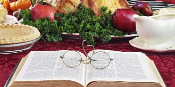 Open Bible with glasses lying on a holiday dinner table with prepared turkey and fixings in background.